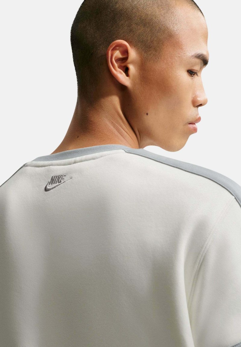 Perfil lateral de un hombre con el cabello corto que lleva una sudadera blanca de Nike con ribetes grises y el logo de Nike en la parte superior de la espalda.