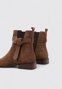 Bottes chelsea en suede marron avec un petit talon bloc, des panneaux latéraux élastiques et un détail de boucle décoratif sur le côté extérieur.