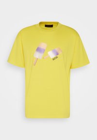 Camiseta de algodón amarilla con un gráfico de dos polos en tonos de púrpura y blanco. El logo de la marca "MCM" está incluido cerca del diseño.