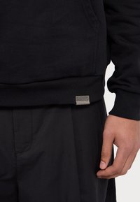 Czarna bluza z teksturowanego materiału, z prążkowanymi mankietami i końcówką. Posiada małą metaliczną tabliczkę z logo na dolnej lewej stronie.