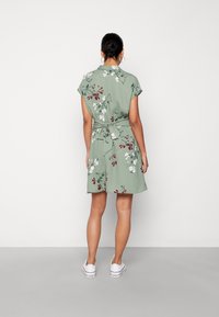 Kort grön klänning med blommönster i vitt och vinrött. Har krage, korta ärmar och ett knytband i midjan. Bärs med vita skor.