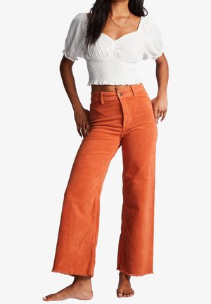 Oranje corduroy wijde broek met een rafelige zoom, gecombineerd met een witte gesmokte crop top. De stof heeft een geribbelde textuur.