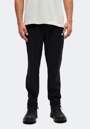 Mann trägt schwarze Freizeithose mit kleinem weißem Logo am Oberschenkel und schwarze Wanderschuhe vor einfachem Hintergrund.