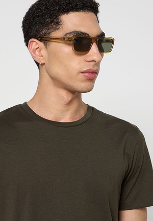 Giovane uomo con t-shirt verde scuro e occhiali da sole marroni trasparenti, volto verso destra su sfondo chiaro uniforme.