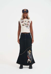 Falda larga de mezclilla negra con un gráfico de águila y rosa, que presenta un dobladillo curvado y detalles de flecos. Combinada con un top corto gráfico blanco y una gorra azul.