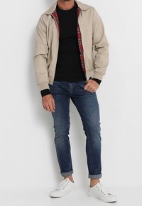 Veste bomber beige avec une doublure à carreaux rouges, t-shirt noir, jeans en denim bleu et baskets blanches ; possède des poignets et un col côtelés.