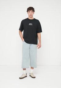 Μαύρο βαμβακερό T-shirt με το λευκό τύπωμα "MICKEY", συνδυασμένο με ανοιχτό μπλε φαρδιά τζιν σορτς και μπεζ αθλητικά παπούτσια. Φαίνονται λευκές κάλτσες.