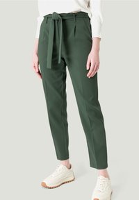 Pantalons ajustés verts avec une ceinture nouée à la taille, comportant des plis et une coupe slim. Associés à des baskets beiges sur un fond neutre.