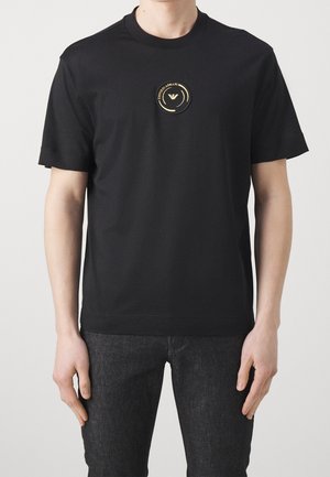 Homme portant un t-shirt noir à manches courtes avec un petit écusson rond du logo Emporio Armani en or au centre de la poitrine.