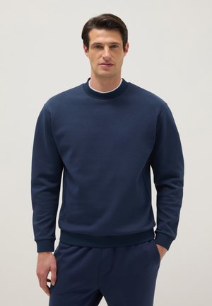 Hombre con cabello oscuro que lleva una sudadera azul marino y pantalones a juego, de pie con una mano en el bolsillo contra un fondo liso.