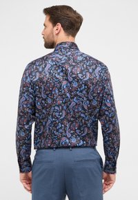 Mann mit dem Rücken zur Kamera, der ein schwarzes langärmliges Hemd mit blauem und rotem floralem Paisleymuster sowie blaue Hosen trägt.