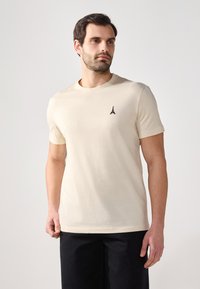 Beige bomull t-skjorte med korte ermer og rund hals. Har et lite svart brodert detalj av Eiffeltårnet på brystet.