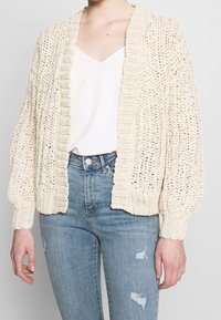 Haak cardigan in roomkleur, met een geribbelde kraag en manchetten, open voorkant en een losse pasvorm. Gecombineerd met lichtblauwe, versleten jeans.