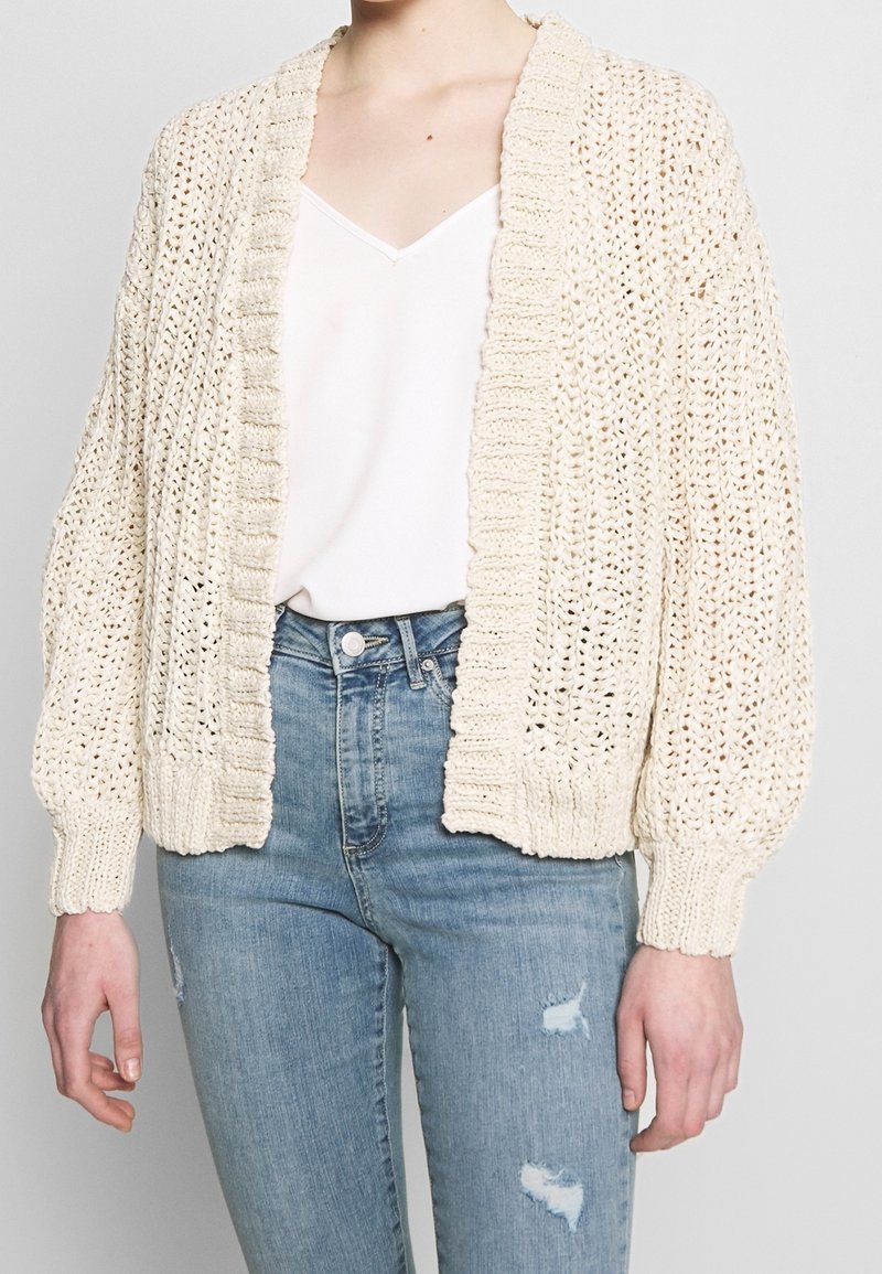 Haak cardigan in roomkleur, met een geribbelde kraag en manchetten, open voorkant en een losse pasvorm. Gecombineerd met lichtblauwe, versleten jeans.