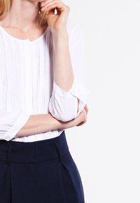 White blouse with pleated front detail, long sleeves, and button cuffs, paired with dark blue tailored trousers. Clean design, lightweight fabric.