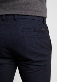 Pantalon tissé bleu marine avec une finition texturée, comportant une poche arrière et un accent en cuir marron près de la ceinture.