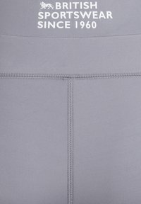 Leggings sportivi grigi caratterizzati da un tessuto liscio e elastico, cuciture piatte e testo stampato in bianco che recita "ABBIGLIAMENTO SPORTIVO BRITANNICO DAL 1960."