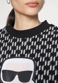 Schwarzer Pullover mit einem sich wiederholenden 'K'-Logo-Muster, runder Halslinie und einer zentralen Grafik einer Figur mit Sonnenbrille.