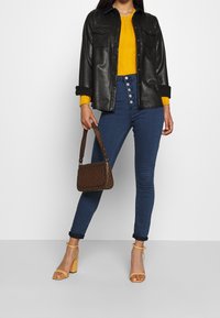 Schwarze Lederjacke, senfgelber Rippstrickpullover, hochtaillierte dunkelblaue Jeans mit Knopfdetails, braune gemusterte Handtasche und beige Absätze.