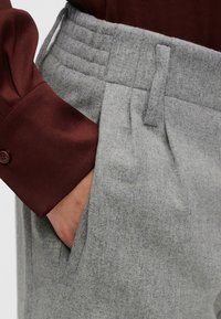 Pantalon en mélange de laine gris avec une ceinture texturée, doté de poches latérales. Un haut bordeaux est partiellement visible à la manche.