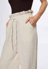 Pantalons larges beiges en tissu texturé, avec une ceinture élastique froncée et des poches latérales ; détail de cordon de serrage à l'avant.