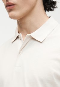 Κοντινό πλάνο ενός ατόμου που φοράει ανοιχτό μπεζ πόλο με γιακά και δύο κουμπιά κλεισμένα, εμφανίζοντας τον λαιμό και το κάτω μέρος του προσώπου.