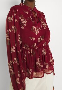 Blouse florale bordeaux en tissu transparent, manches longues, fermeture avec noeud au col, et taille cintrée. Motif floral rose et vert.
