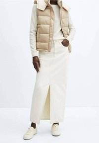 Beiger Puffer-Weste, cremfarbener Pullover und langer, off-white Jeansrock mit einem Schlitz vorne. Kombiniert mit hellen Sneakers. Einfaches Design.