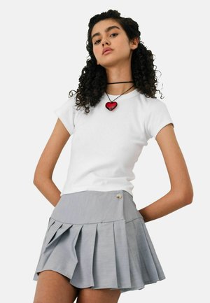 Joven con cabello rizado que lleva una camiseta blanca ajustada, falda plisada gris claro y un collar con colgante en forma de corazón rojo.