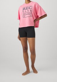 Růžové zkrácené tričko s grafickým potiskem a textem, spojené s černými šortkami. Měkký bavlněný materiál s uvolněným střihem.