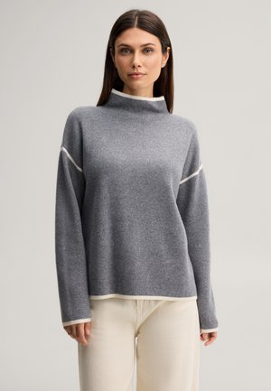 Femme portant un pull gris à manches longues avec bordure blanche et un pantalon beige clair, debout devant un fond uni.