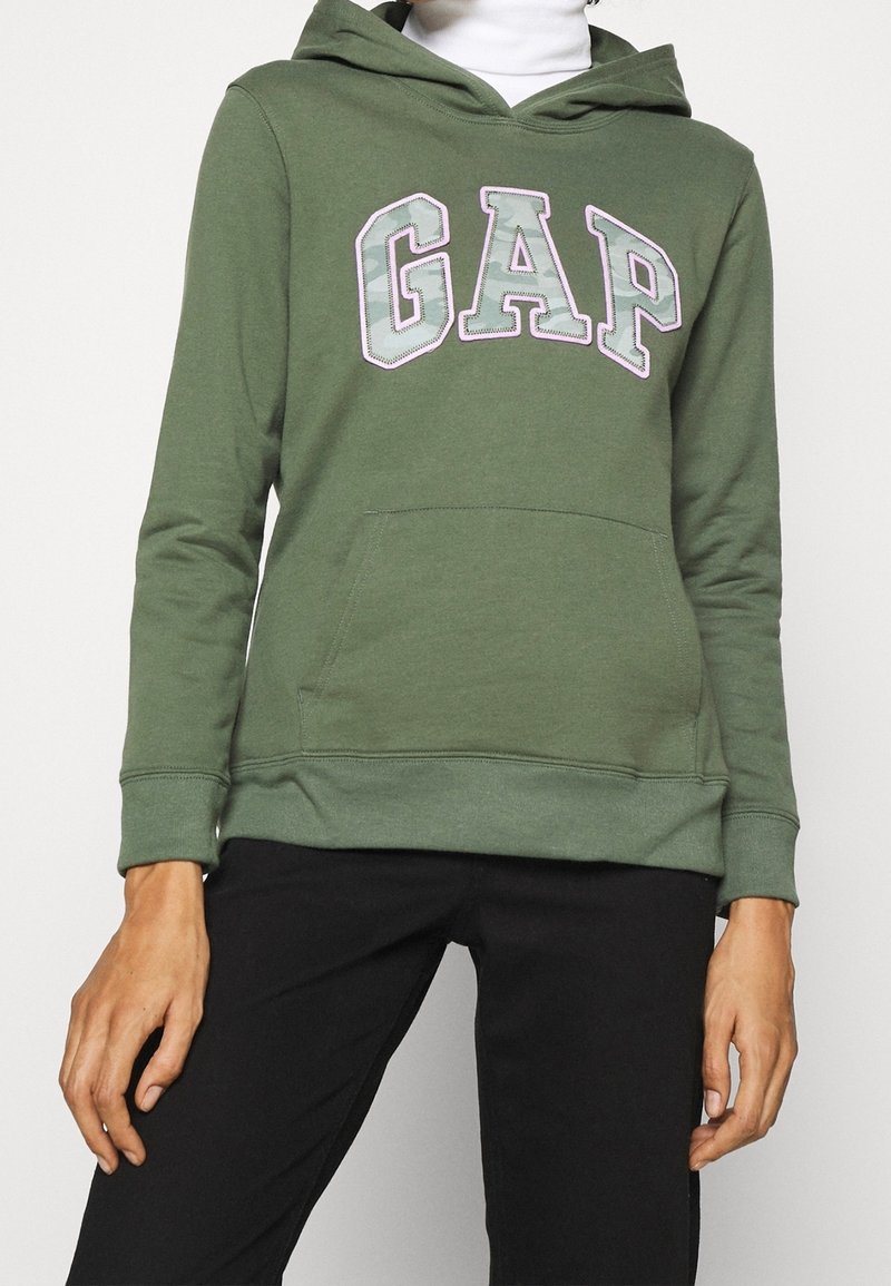 Personne portant un sweat à capuche GAP vert olive avec poche kangourou et pantalon noir, debout devant un fond clair uni.
