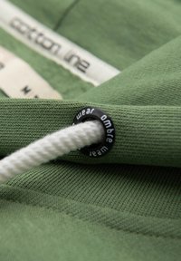 Tissu vert avec un cordon blanc passé à travers un œillet en métal noir marqué "ombre wear" et une étiquette de vêtement floue en arrière-plan.