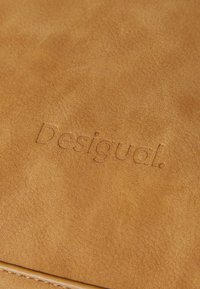 Tan skinnemateriale har et preget "Desigual"-logo, jevn tekstur med minimalt synlige sømmer eller maskinvarekontoer.