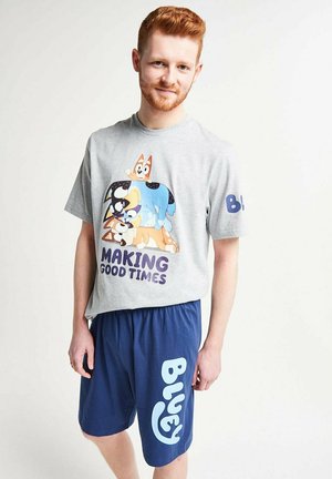Junger Mann trägt ein graues T-Shirt mit gestapelten Cartoon-Hunden und dem Schriftzug "Making Good Times", kombiniert mit marineblauen Shorts mit der Aufschrift "Bluey."