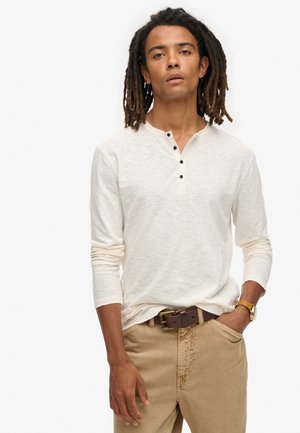 Camisa henley de manga larga, color off-white, con tres botones negros en el cuello. Tejido texturizado, combinada con pantalones marrón claro y un cinturón marrón.