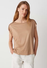 Lichtbruine top met korte mouwen, ontspannen pasvorm en ronde halslijn. Zachte textuur, minimale stiksels en schouderaccenten. Draag met witte broek.