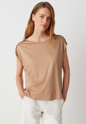 Haut marron clair à manches courtes avec une coupe décontractée et un col rond. Texture douce, couture minimale et accents aux épaules. Porté avec un pantalon blanc.
