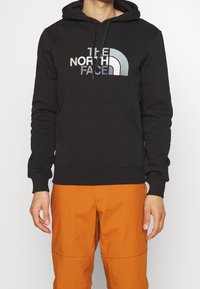 Uomo che indossa una felpa nera con cappuccio con un grande logo "THE NORTH FACE" e pantaloni arancioni, in piedi su uno sfondo bianco semplice.