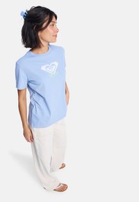 Světle modré bavlněné tričko s bílým logem srdce a nápisem "ROXY" v zelené barvě. V kombinaci s volnými, světlými kalhotami a sandály.