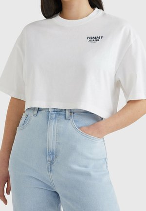Nainen pukeutunut valkoiseen lyhyeen Tommy Jeans -t-paitaan ja vaaleansinisiin korkeavyötäröisiin farkkuihin, yksi käsi taskussa.