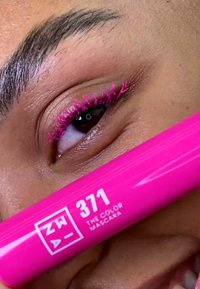 Tube de mascara rose vif étiqueté "371 THE COLOR MASCARA" tenu près de l'œil avec des cils roses et pointus. Peau lisse et œil marron foncé visible.