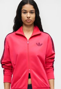 Jeune femme aux longs cheveux foncés portant une veste zippée Adidas rose vif avec des bandes noires sur les manches et un logo sur la poitrine.