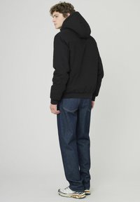 Veste noire rembourrée avec capuche ajustable, poignets élastiques et base. Associée à un jean en denim bleu foncé et à des chaussures de sport blanches.