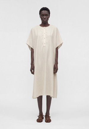 Žena stoji okrenuta naprijed, nosi labavu, bež haljinu do koljena s vertikalnim prugama i gumbima, uparenu s smeđim sandalama.