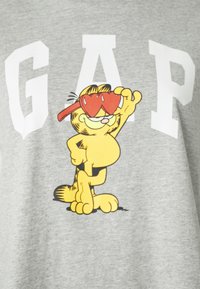 Camisa gris de manga corta con un personaje de gato de dibujos animados que lleva gafas de sol en forma de corazón, con letras blancas en negrita que dicen "GAP."