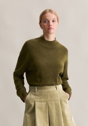 Olivgrüner Strickpullover mit hohem Kragen und langen Ärmeln, kombiniert mit einem hellgrünen Cordrock mit Falten und Taschen.