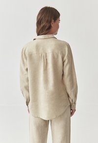 Vaalean beige pellavapaita, jossa on pitkät hihat, kauluskangas ja kaareva helma. Sisältää nappisuljettavat hihansuut ja hienovaraisen tekstuurin.