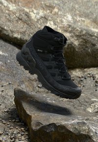 Black hiking boots with a high ankle, made from synthetic material, featuring a textured pattern and Gore-Tex branding, displayed on rocky terrain.