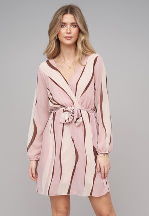 Kvinde med langt blondt hår iført en lyserød og brun stribet langærmet slå-om-kjole bundet i taljen, stående mod en ensfarvet grå baggrund.